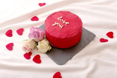 Round red bento cake, hearts and roses lie on white bed in daylight Stock Photos