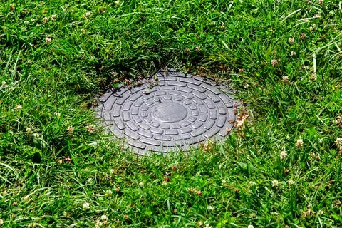 Round sewer manhole with a pattern on a green lawn on a sunny summer day. Stock Photos