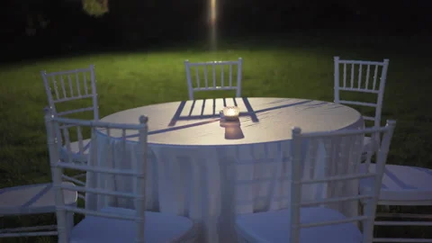Round table covered with tablecloth and white chairs in the garden at night Stock Footage 231426527