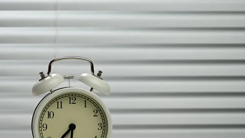 Round white alarm clock strikes a bell against the backdrop of blinds, half past Stock Footage 162040980
