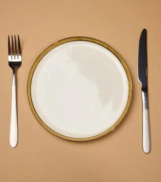 Round white empty ceramic plate and knife with fork on brown background, ta.. Stock Photos