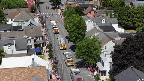 Row of fire trucks drives parade route down tree-lined street in small American 库存影片 340968688
