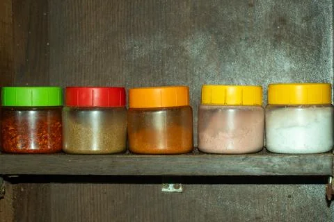 Row of five colorful plastic spice jars on dark wooden shelf close. Stock Photos