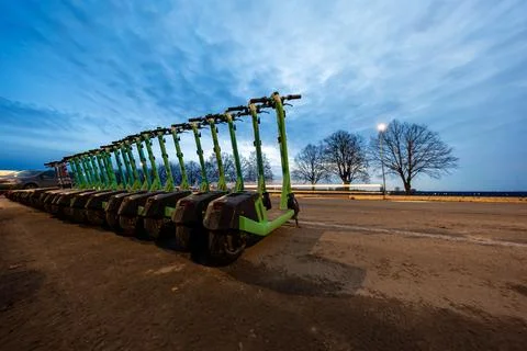 Row of lime e scooters at dusk beside a waterfront road and trees Stock Photos