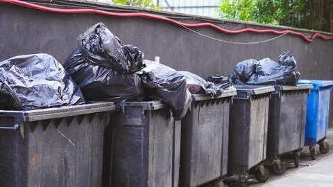 Row of overflowing garbage bins with black plastic bags on street side Stock Photos
