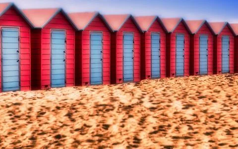Row of vibrant red beach huts with blue doors set against a sandy beach and.. Stock Photos