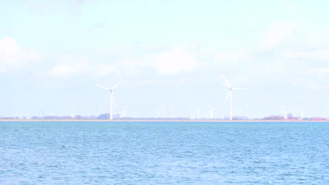 A row of wind turbines rises along the distant shoreline above calm water. Stock Footage 322689794