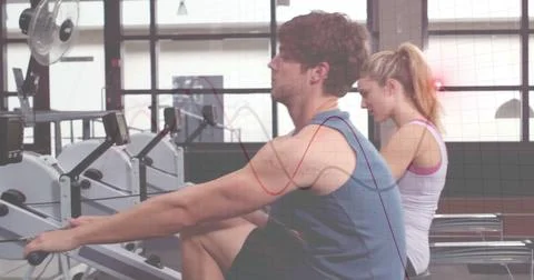 Rowing partners pushing handle bars with foot rests on rowers at fitness center, Stock Photos