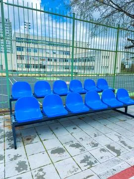 Rows of blue chairs sit empty beside a sports field. A school building stands Stock Photos