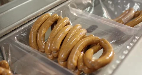 Rows of brown sausage lying in transparent plastic trays on a packaging line in Vídeo Stock 339984701