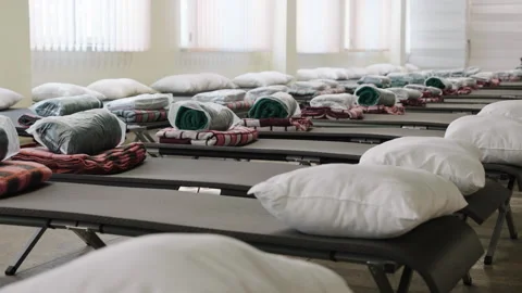 Rows of cots with blankets and pillows in a shelter space during a community Видео 345030584