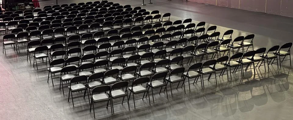 Rows of empty black folding chairs in a large event hall with dark floor conc Stock-Fotos