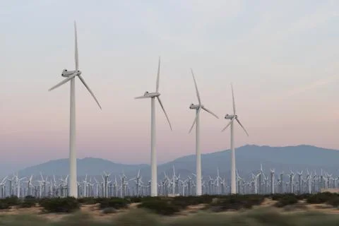 Rows of wind generators set against soft pink, orange sunset sky, mountains in Stock Photos