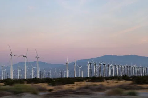 Rows of wind generators set against soft pink, orange sunset sky, mountains in Stock Photos