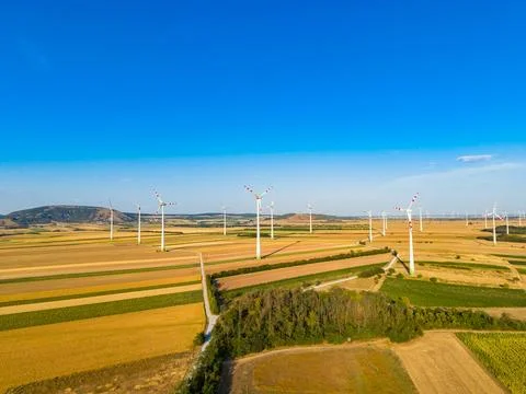 Rows of wind turbines turn slowly on a bright day in a flat area with farmlan Stock Photos