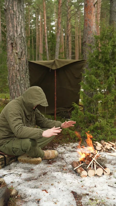 A rugged man warms his hands while sitting by the fire in a coniferous forest in Video stock 308058164