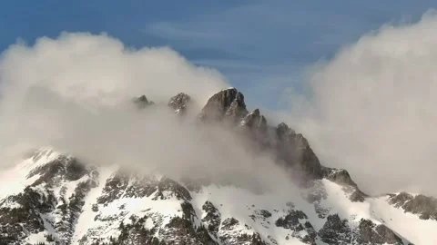 Rugged Snowy Mountain Peak surrounded by fog and Clouds. Sun and blue sky. Stock Footage 282233306
