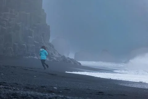 Runner in blue jacket on black sand beach Iceland with misty basalt cliffs Foto stock