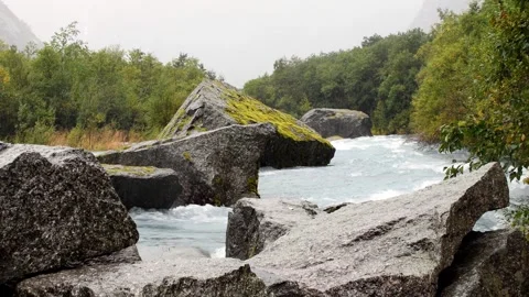 A rushing river flows past mossy rocks and lush green trees under overcast sky Video stock 322833227