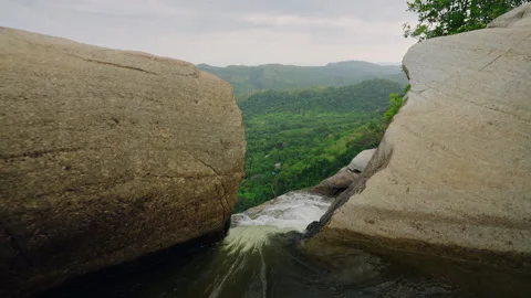 Rushing water flows from river into waterfall over rocks, green hills surround Stock Footage 274227447