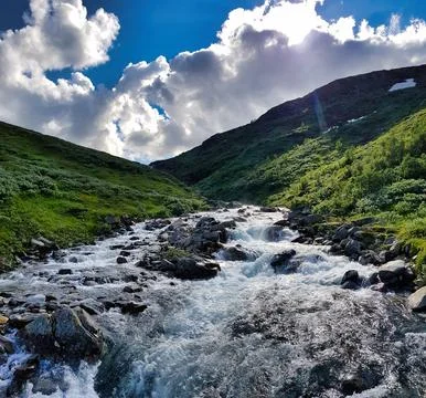 Rushing water flows through rocks in a green valley under bright clouds Stock Photos