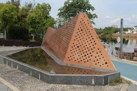 Rust colored triangular metal sculpture with perforated pattern beside a wa.. Stock Photos
