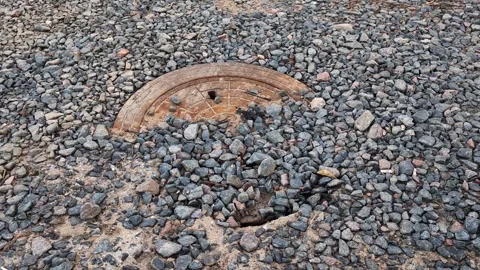 Rusted circular metal drainage cover partially submerged in grey gravel and Stock Footage 344845786