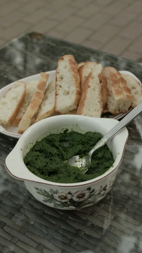 Rustic bowl of green herb dip with sliced bread on glass table in casual outdoor Stock Footage 345268533