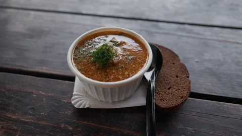 Rustic bowl of hot soup topped with fresh dill beside rye bread on weathered 스톡 동영상 344771107