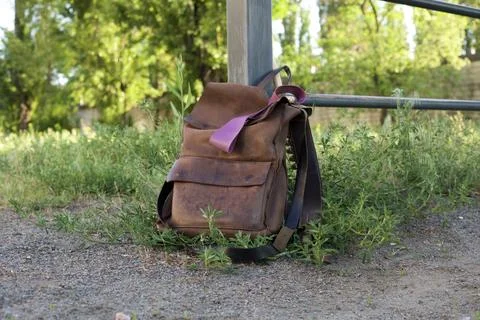 Rustic brown leather backpack resting against a metal fence post outdoors Фото