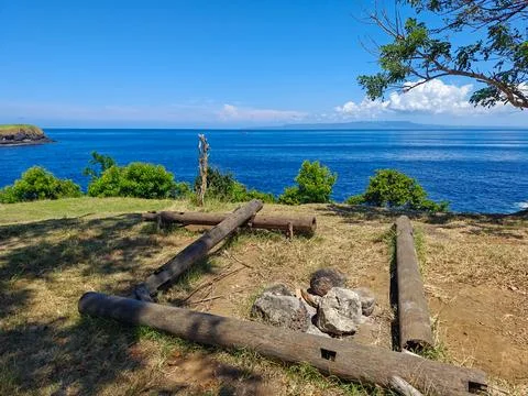 Rustic campfire pit with wooden logs on grassy cliff overlooking blue ocean Foto stock