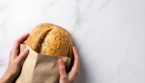 Rustic grain wheat bread in a paper bag in female hands on white marble. Organic Stock Photos