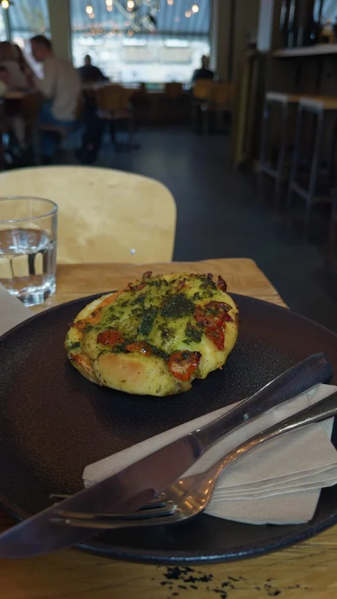 Rustic herb topped baked bread on dark plate beside cutlery, napkin and water Stock Footage 345124474
