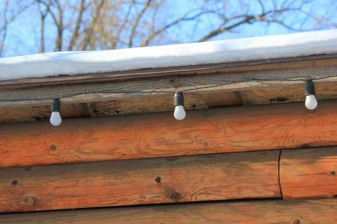 Rustic log cabin wall with a snow-covered roof and a hanging garland of light Stock Photos