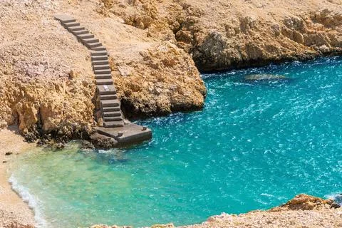 Rustic stone steps leading to vibrant turquoise sea on rocky coastline Stock Photos