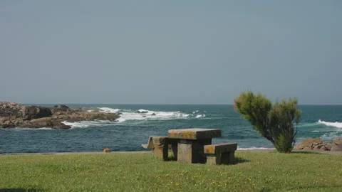 Rustic stone table and bench set on a green, flower-speckled grassy cliff Видео 344203764