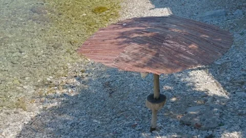 A rustic straw beach umbrella stands on a pebbly shoreline next to clear Stock Footage 346677260