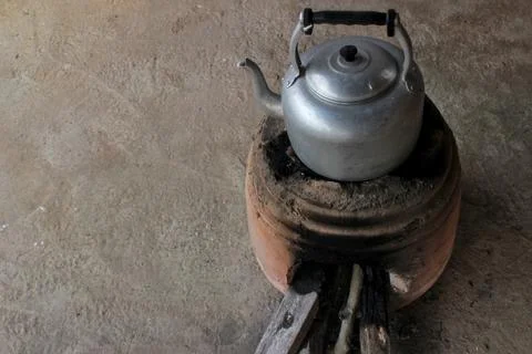 A rustic-style silver aluminum kettle sits atop an earthen stove. Stock Photos