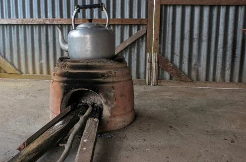 A rustic-style silver aluminum kettle sits atop an earthen stove. Stock Photos