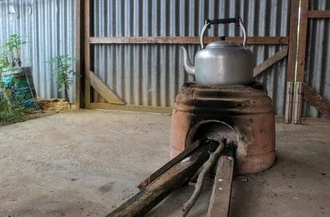 A rustic-style silver aluminum kettle sits atop an earthen stove. Stock Photos
