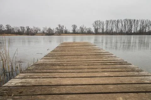 A rustic wooden dock extends into a partially frozen lake under a grey winter Stock Photos