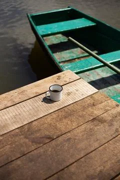 A rustic wooden dock by the water features a single enamel mug of coffee, o.. Stock Photos