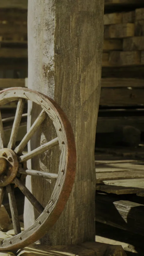 Rustic wooden wheel resting against an old post in a sunlit barn Stock Footage 331829399