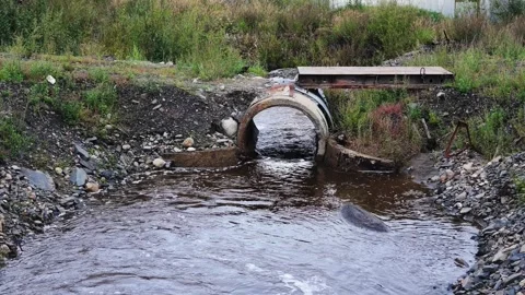 Rusty arched pipe discharges murky water into rocky stream with weathered Stock Footage 344317958