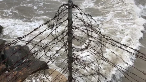 Rusty barbed wire fence on a rocky shore with rough sea waves crashing Stock Footage 336246512