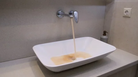 Rusty brown water flowing from chrome faucet into white ceramic sink in modern Stock Footage 345341792