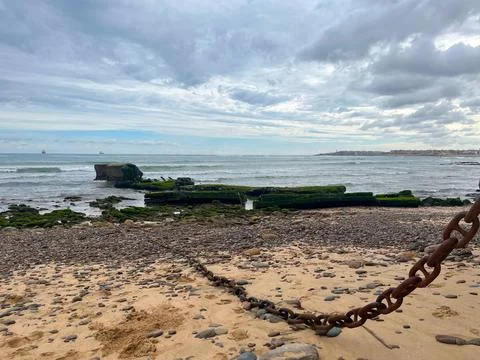 Rusty chain lying on a sandy beach with concrete ruins in the water Stock Photos