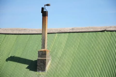 Rusty chimney shadows an aged green corrugated metal roof under blue sky Stock Photos