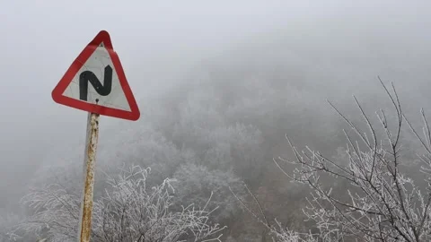 Rusty cracked road sign with sharp curve symbol stands in foggy winter mountain Stock Footage 318685422