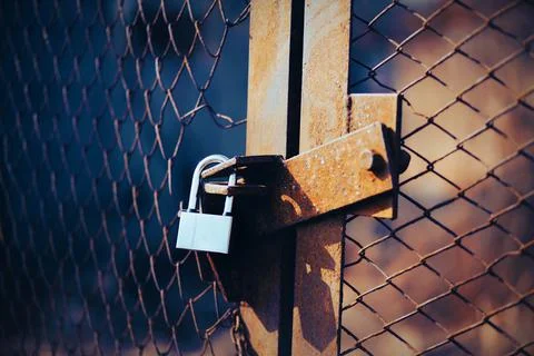 A rusty old gate with a torn metal grid is closed by a strong padlock on a su Stock Photos